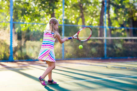 Child playing tennis on outdoor court. Little girl with tennis racket and ball in sport club. Active exercise for kids. Summer activities for children. Training for young kid. Child learning to play.の写真素材
