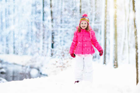 Child playing with snow in winter. Little girl in colorful jacket and knitted hat catching snowflakes in winter park on Christmas. Kids play and jump in snowy forest. Children catch snow flakesの写真素材