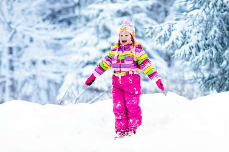 Child playing with snow in winter. Little girl in colorful jacket and knitted hat catching snowflakes in winter park on Christmas. Kids play and jump in snowy forest. Children catch snow flakesの写真素材