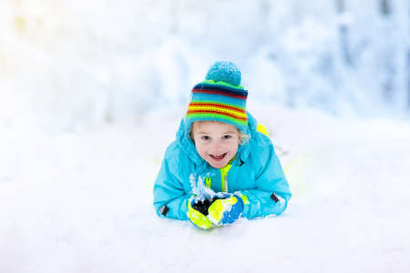 Child playing with snow in winter. Little boy in colorful jacket and knitted hat catching snowflakes in winter park on Christmas. Kids play and jump in snowy forest. Children catch snow flakesの写真素材