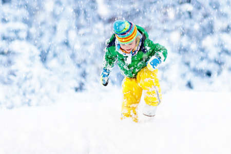 Child playing with snow in winter. Little boy in colorful jacket and knitted hat catching snowflakes in winter park on Christmas. Kids play and jump in snowy forest. Children catch snow flakesの写真素材