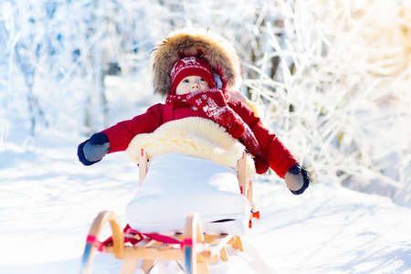Sled and snow fun for kids. Baby sledding in snowy winter park. Little boy in warm red jacket and knitted hat sitting in sheepskin footmuff. Kid on sleigh. Child on sledge. Family Christmas vacation.の写真素材