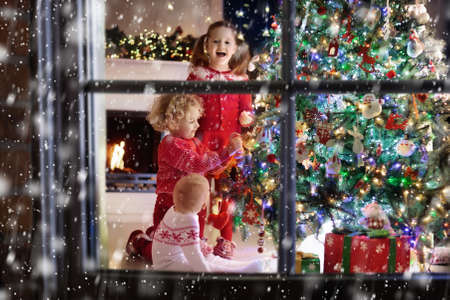 Children at Christmas tree and fireplace on Xmas eve. Family with kids celebrating Christmas at home. Boy and girl in matching sweater decorating xmas tree and opening presents. Holiday gifts for kid.の写真素材