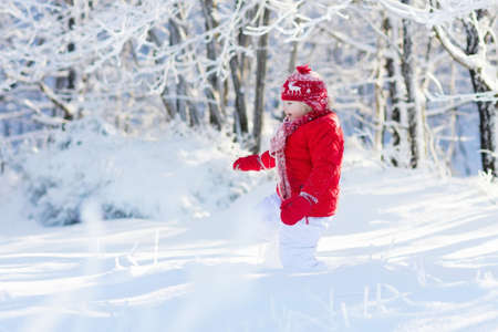 Child playing with snow in winter. Little boy in red jacket and knitted hat catching snowflakes in...