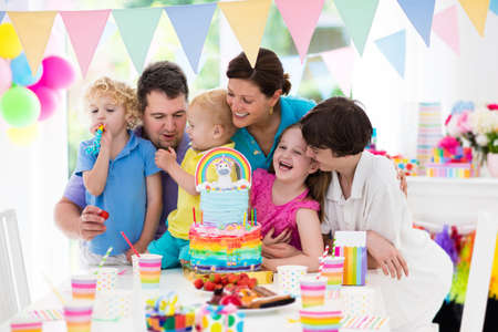 Kids birthday party with unicorn rainbow cake for little girl. Family celebrating child birthday. Parents and children blowing candles at baby cake. Balloons and colorful pastel banner decoration.の写真素材