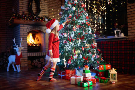 Child at Christmas tree and fireplace on Xmas eve. Family with kids celebrating Christmas at home. Little girl decorating xmas tree and opening presents. Gifts for winter holidays at fire place.の写真素材
