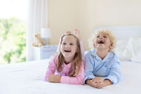 Kids playing in parents bed. Children wake up in sunny white bedroom. Boy and girl play in matching pajamas. Sleepwear and bedding for child and baby. Nursery interior for toddler kid. Family morning.の写真素材