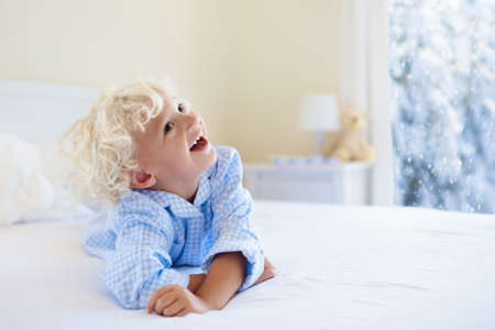 Kids playing in parents bed in winter. Children wake up in white bedroom by snow. Little boy in blue pajamas. Christmas sleepwear and bedding for child and baby. Family morning on snowy cold Xmas day.の写真素材