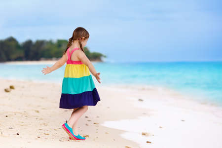 Kid playing on tropical beach. Child running and dancing at sea shore. Kids play on ocean coast. Water and sand fun for children. Summer dress fashion for little girl. Family summer beach vacation.の写真素材