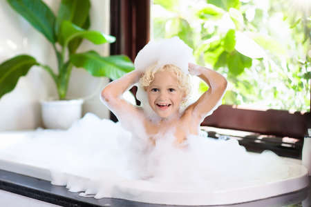 Little child taking bubble bath in beautiful bathroom with big garden view window. Kids hygiene....