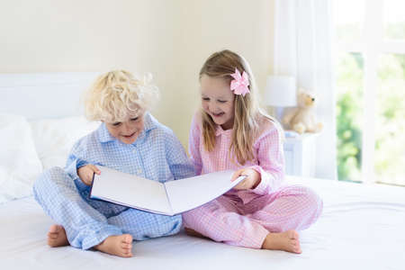 Kids playing in parents bed. Children wake up in sunny white bedroom. Boy and girl play in matching pajamas. Sleepwear and bedding for child and baby. Nursery interior for toddler kid. Family morning.の写真素材