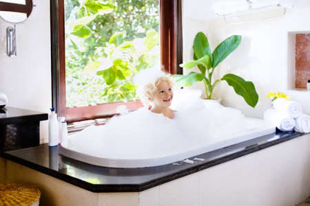 Little child taking bubble bath in beautiful bathroom with big garden view window. Kids hygiene. Shampoo, hair treatment and soap for children. Kid bathing in large tub. Baby boy with foam in hair.の写真素材