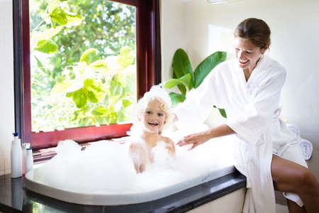Little child taking a bubble bath in a beautiful bathroom with a big garden view window. Mother...
