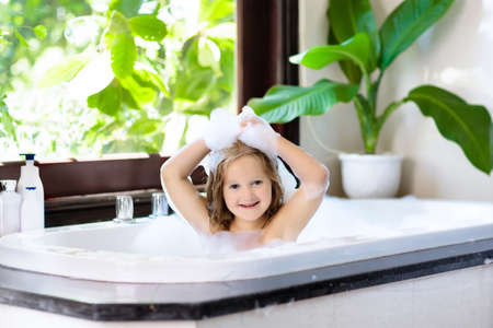 Little child taking bubble bath in beautiful bathroom with big garden view window. Kids hygiene. Shampoo, hair treatment and soap for children. Kid bathing in large tub. Girl with foam in hair.の写真素材