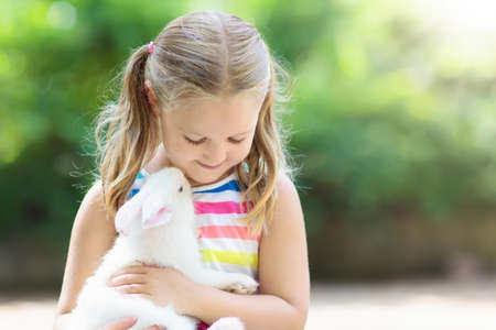 Child playing with white rabbit. Little girl feeding and petting white bunny. Easter celebration. Egg hunt with kid and pet animal. Children and animals. Kids take care of pets. Spring Easter garden.の写真素材