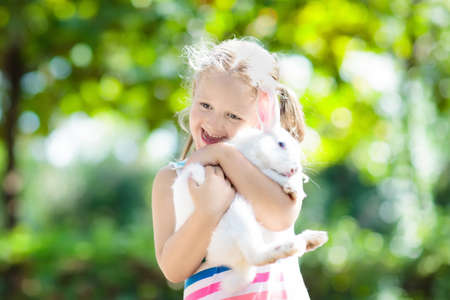 Child playing with white rabbit. Little girl feeding and petting white bunny. Easter celebration. Egg hunt with kid and pet animal. Children and animals. Kids take care of pets. Spring Easter garden.の写真素材
