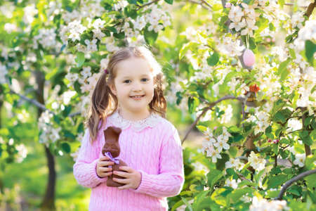 Kids with bunny ears on Easter egg hunt in blooming cherry blossom garden. Little girl eating chocolate rabbit. Spring flowers and eggs basket in fruit orchard. Children with Easter candy and sweets.の写真素材