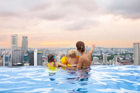 Family swimming in roof top outdoor pool on family vacation in Singapore. City skyline from infinity pool in luxury hotel. Kids swim and enjoy skyscraper view in Asia. Travel with young children.の写真素材