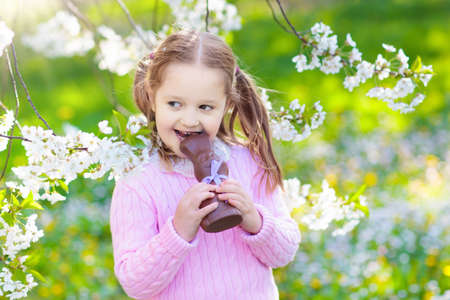 Kids with bunny ears on Easter egg hunt in blooming cherry blossom garden. Little girl eating chocolate rabbit. Spring flowers and eggs basket in fruit orchard. Children with Easter candy and sweets.の写真素材