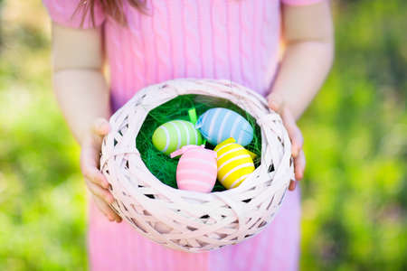 Easter egg hunt. Little girl with eggs basket. Kids searching for sweets and chocolate on Easter morning in garden. Child with spring pastel decoration.  Kid in pink dress with painted eggs.の写真素材