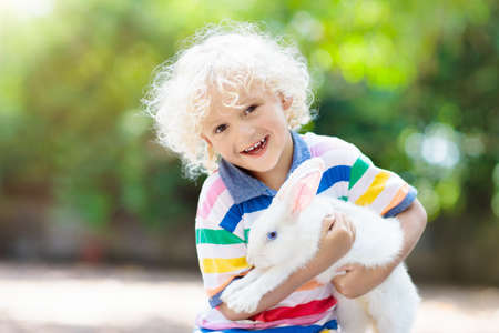 Child playing with white rabbit. Little boy feeding and petting white bunny. Easter celebration. Egg hunt with kid and pet animal. Children and animals. Kids take care of pets. Spring Easter garden.の写真素材