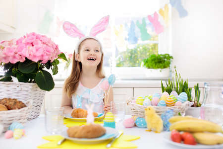 Family Easter breakfast. Child with bunny ears at decorated table with eggs basket, chick and rabbit on Easter morning. Egg hunt and festive meal for kids. Spring home decoration. Girl with dyed eggs.の写真素材
