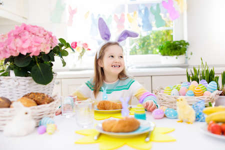 Family Easter breakfast. Child with bunny ears at decorated table with eggs basket, chick and rabbit on Easter morning. Egg hunt and festive meal for kids. Spring home decoration. Girl with dyed eggs.の写真素材