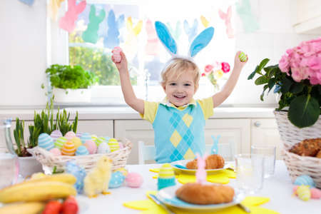 Family Easter breakfast. Child with bunny ears at decorated table with eggs basket, chick and rabbit on Easter morning. Egg hunt and festive meal for kids. Spring home decoration. Boy with dyed eggs.の写真素材