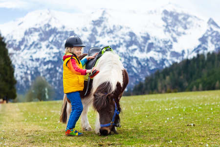 Kids riding pony in the Alps mountains. Family spring vacation on horse ranch in Austria, Tirol. Children ride horses. Kid taking care of animal. Child and pet. Little boy in saddle on pony.の写真素材