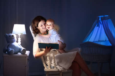 Mother and baby reading a book in dark bedroom. Mom and child read books before bed time. Family in...
