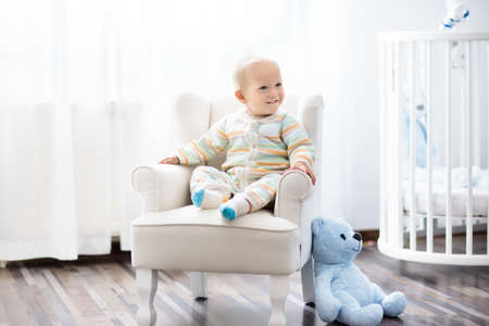 Baby boy in bedroom. Infant child sitting in white chair at round wooden bed. Kids room interior. Children fashion and sleep wear. Kid at nursery crib. Toys for toddler. Knitting for kids.の写真素材