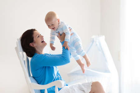 Young mother holding her newborn child. Mom nursing baby. Woman and new born boy in white bedroom with rocking chair and blue crib. Nursery interior. Mother playing with laughing kid. Family at homeの写真素材