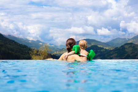 Mother and baby play in outdoor infinity swimming pool of luxury spa alpine resort at sunset in Alps mountains. Spring or summer vacation with children. Family with kids in hot tub with mountain view.の写真素材