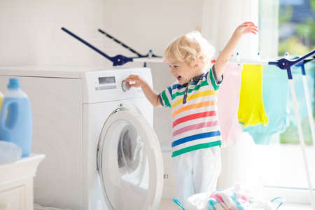 Child in a laundry room with washing machine or tumble dryer. Kid helping with family chores. Modern household devices and washing detergent in white sunny home. Clean washed clothes on drying rack.の写真素材