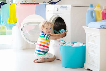 Child in a laundry room with washing machine or tumble dryer. Kid helping with family chores. Modern household devices and washing detergent in white sunny home. Clean washed clothes on drying rack.の写真素材