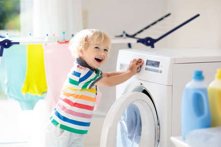 Child in a laundry room with washing machine or tumble dryer. Kid helping with family chores. Modern household devices and washing detergent in white sunny home. Clean washed clothes on drying rack.の写真素材