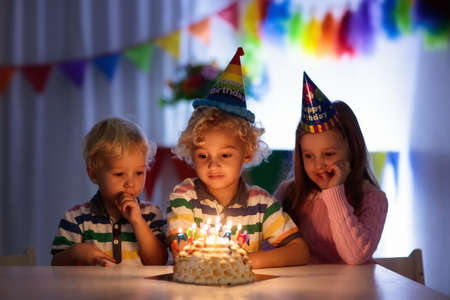 Kids birthday party. Children blow out candles on cake in dark room. Rainbow decoration and table setting for kids event, banner and flag. Girl and boy with birthday presents. Family celebration.の写真素材