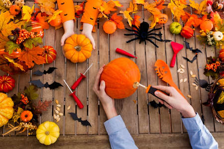 Family carving Halloween pumpkin on wooden table with autumn leaves decoration. Top view flat lay of hands and carved pumpkins. Halloween trick or treat. Seasonal home dÃ©cor.