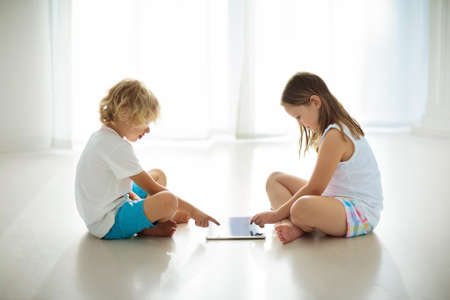 Child with touch screen tablet pc. Kid playing computer game. Little girl and boy in white bedroom...