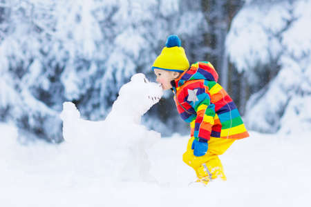 Kid making snowman in snowy winter park. Children play in snow. Baby boy in colorful jacket and hat building snow dinosaur in winter garden after snowfall. Outdoor fun on cold winter day.の写真素材