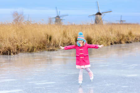 Child ice skating on frozen canal with wind mills and snow in Holland. Little girl with skates on natural ice on cold winter day in the Netherlands. Kids ice skate in snowy Dutch windmill landscape.の写真素材