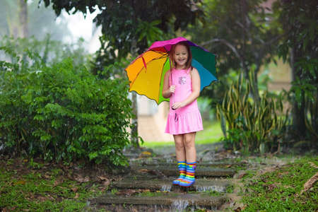 Kid playing out in the rain. Children with umbrella and rain boots play outdoors in heavy rain. Little girl jumping in muddy puddle. Kids fun by rainy autumn weather. Child running in tropical storm.の写真素材