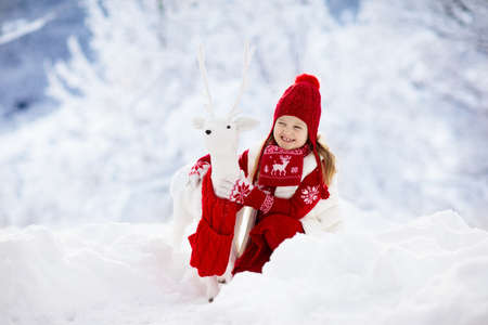 Child playing with reindeer in snow on Christmas vacation. Winter outdoor fun. Kids play in snowy park on Xmas eve. Little girl in knitted sweater, hat, scarf and mittens with Christmas decoration.の写真素材