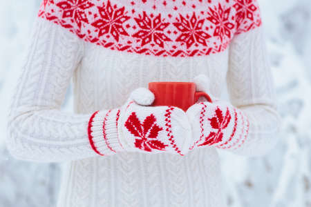 Woman drinking hot chocolate in Christmas morning in snowy garden. Girl in knitted Nordic sweater, hat and mittens holding cup with cocoa. Snow and winter outdoor fun. Glogg mug in female hands.の写真素材
