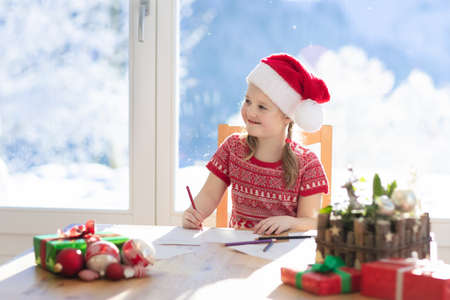 Child writing letter to Santa on Christmas eve. Kids write Xmas present wish list. Little girl sitting in decorated living room with big window on sunny winter day in the mountains, snow outside.の写真素材