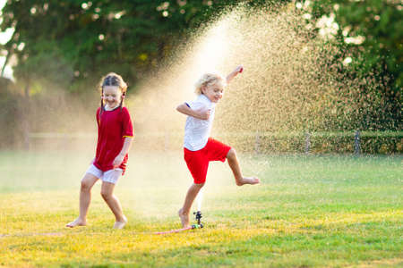 Kids play with water on hot summer day. Children with garden sprinkler. Outdoor fun. Boy and girl run on football field after training under water drops.の写真素材