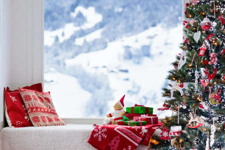 Christmas tree at window with view to snow and Swiss Alps mountains in winter. Decorated living room with Xmas gifts and presents for kids, pillows and toys. Family home seasonal interior decoration.の写真素材