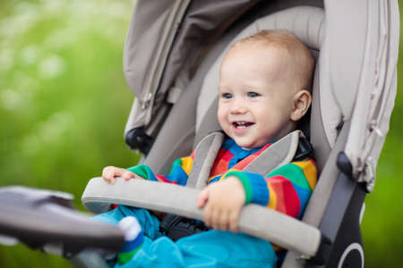 Baby boy in warm colorful knitted jacket sitting in modern stroller on a walk in a park. Child in...
