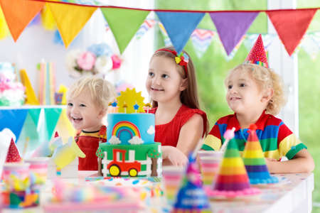 Kids birthday party. Child blowing out candles on colorful cake. Decorated home with rainbow flag banners, balloons, confetti. Farm and transport theme. Little boy celebrating birthday. Party food.の写真素材