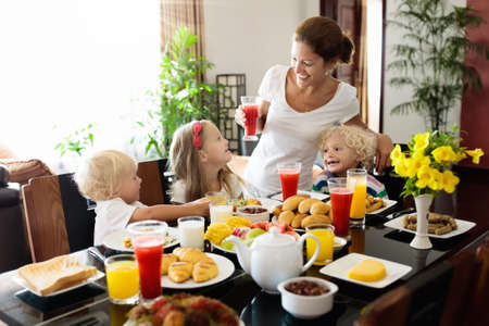 Healthy family breakfast at home. Mother and kids eating tropical fruit, toast bread, cheese and sausage. Children drink fresh pressed juice on sunny morning. Mom, boy, girl and baby eat breakfast.